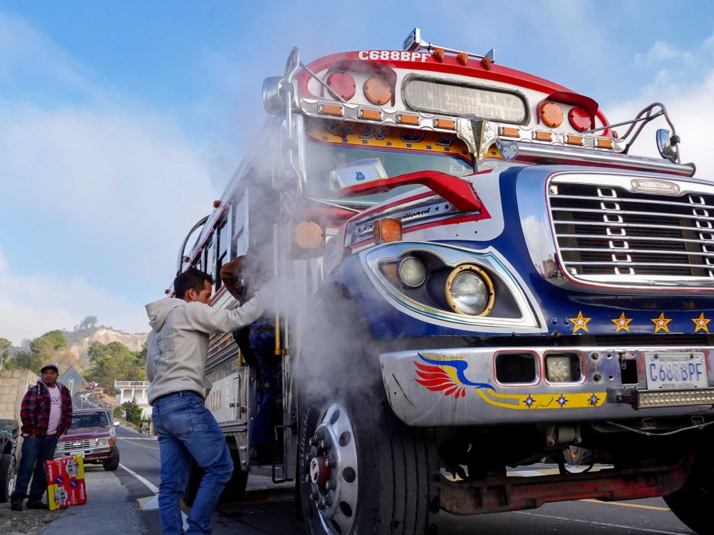 Driver repairing the engine of a steaming chicken bus on the roadside.
